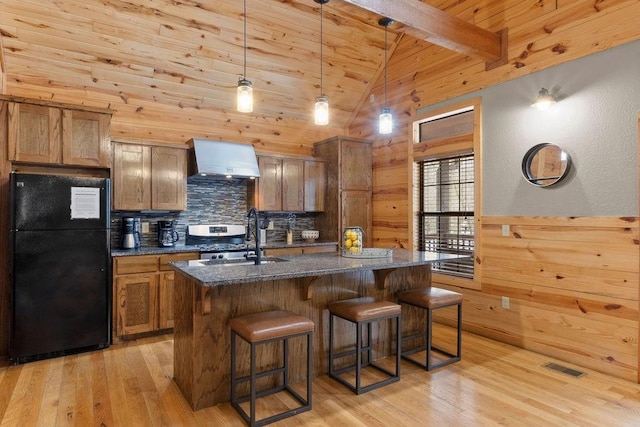 kitchen with an island with sink, black fridge, wall chimney exhaust hood, beam ceiling, and sink