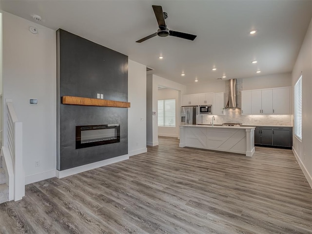 kitchen with white cabinetry, an island with sink, stainless steel appliances, backsplash, and wall chimney range hood