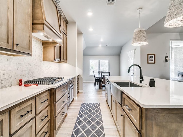 kitchen with a center island with sink, custom exhaust hood, decorative backsplash, sink, and hanging light fixtures