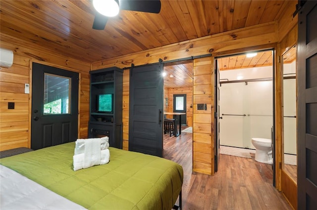 bedroom featuring connected bathroom, a barn door, wood walls, wood-type flooring, and ceiling fan