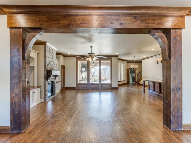 unfurnished living room featuring dark hardwood / wood-style flooring, a stone fireplace, ceiling fan with notable chandelier, and ornamental molding