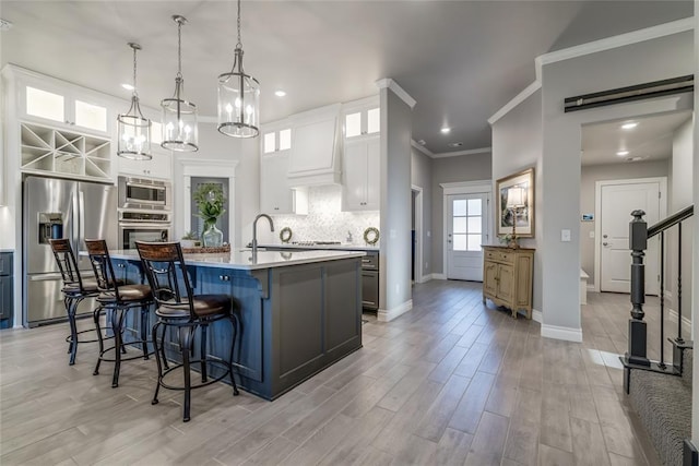 kitchen with appliances with stainless steel finishes, an island with sink, white cabinets, decorative light fixtures, and custom exhaust hood