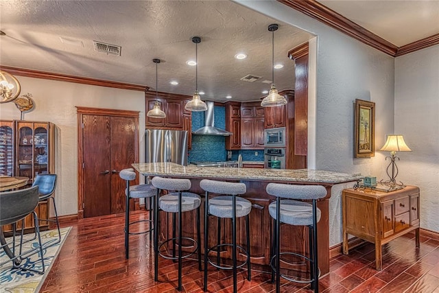 kitchen featuring a breakfast bar area, appliances with stainless steel finishes, ornamental molding, wall chimney exhaust hood, and decorative light fixtures