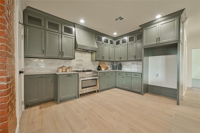 kitchen featuring wall chimney range hood, backsplash, light hardwood / wood-style floors, stainless steel stove, and green cabinetry