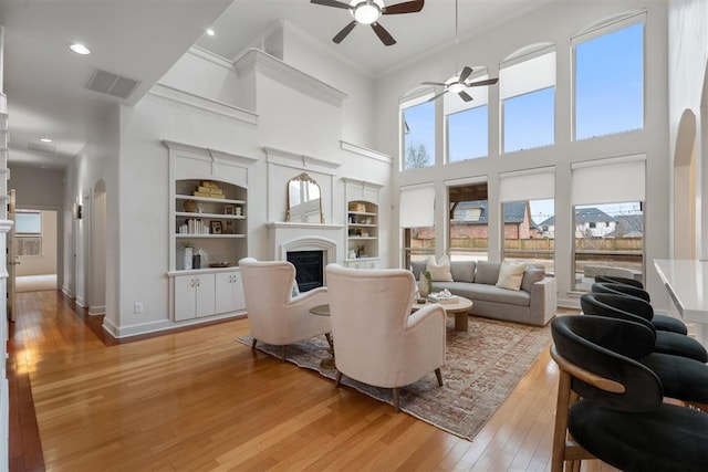 living room with ceiling fan, a towering ceiling, crown molding, light wood-type flooring, and built in shelves