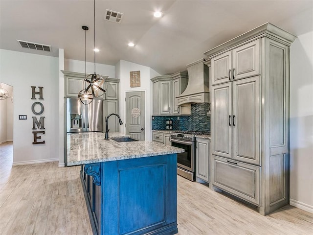 kitchen with appliances with stainless steel finishes, wall chimney exhaust hood, sink, light stone counters, and a center island with sink