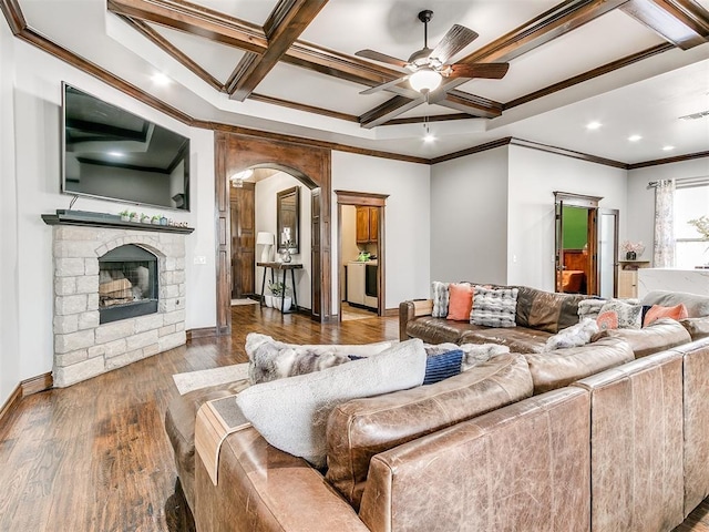 living room featuring a stone fireplace, coffered ceiling, crown molding, ceiling fan, and dark wood-type flooring