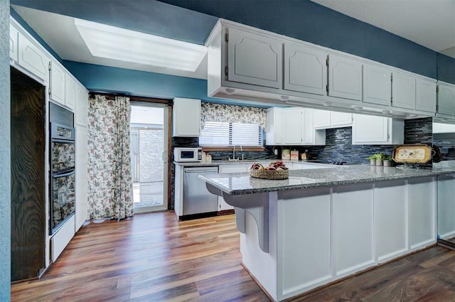 kitchen featuring a peninsula, wood finished floors, white microwave, white cabinets, and dishwasher
