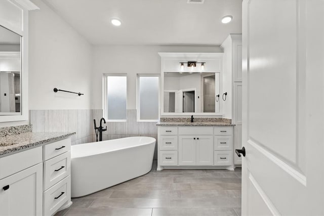 full bathroom featuring a wainscoted wall, a soaking tub, two vanities, a sink, and tile walls