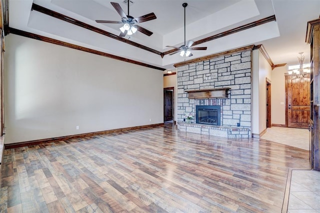 unfurnished living room with a stone fireplace, light hardwood / wood-style floors, ornamental molding, a tray ceiling, and ceiling fan with notable chandelier