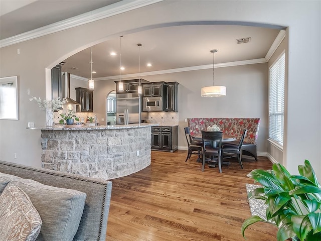 bar featuring wall chimney range hood, crown molding, hanging light fixtures, built in appliances, and decorative backsplash