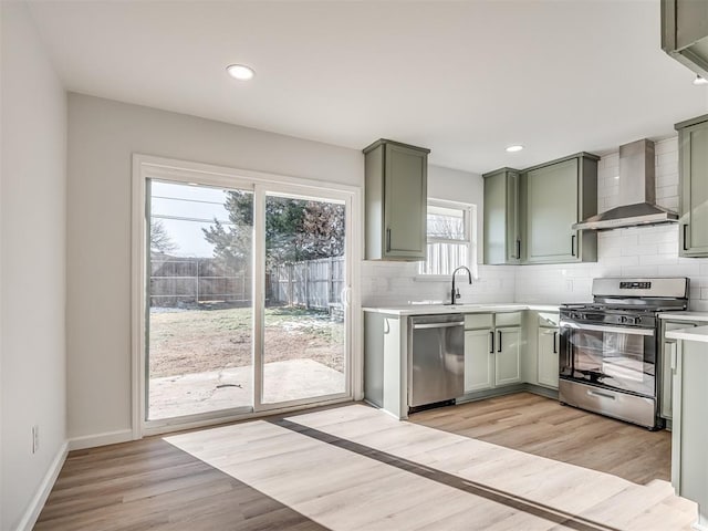 kitchen with green cabinets, light wood-type flooring, backsplash, wall chimney range hood, and appliances with stainless steel finishes