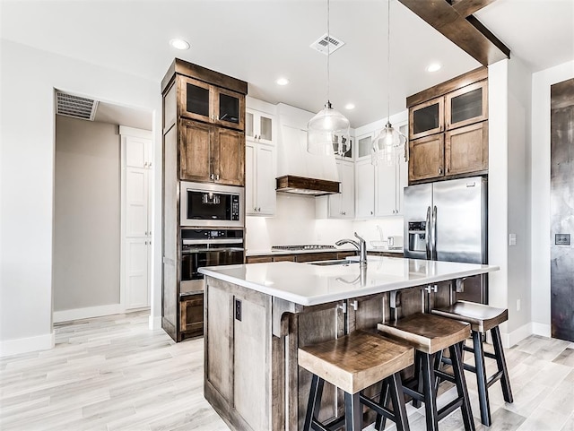 kitchen featuring premium range hood, a center island with sink, appliances with stainless steel finishes, light wood-type flooring, and white cabinets