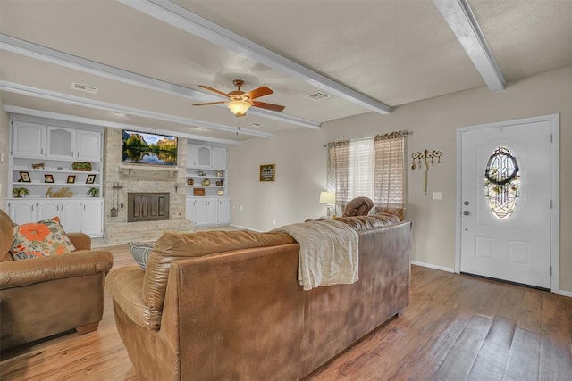 living room featuring a stone fireplace, light wood-type flooring, beamed ceiling, ceiling fan, and built in features