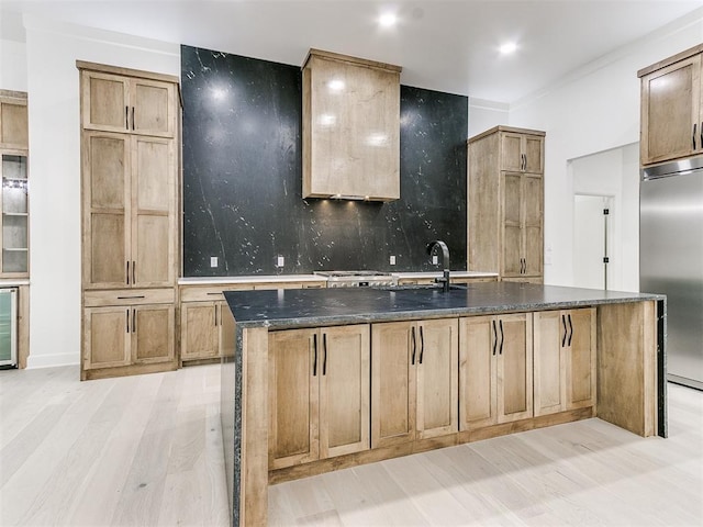 kitchen featuring built in fridge, light hardwood / wood-style flooring, decorative backsplash, dark stone counters, and a center island with sink