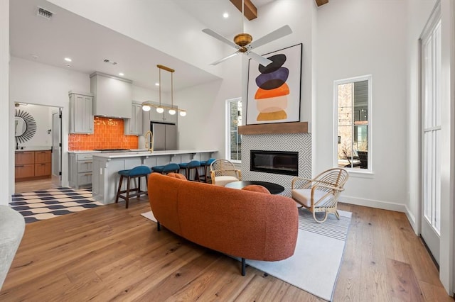 living room with light hardwood / wood-style floors, a towering ceiling, beam ceiling, a tiled fireplace, and ceiling fan