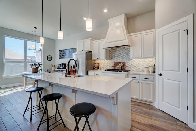 kitchen with decorative light fixtures, a breakfast bar, custom range hood, a kitchen island with sink, and white cabinets