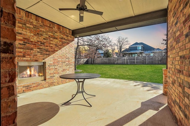 patio terrace at dusk featuring an outdoor brick fireplace, ceiling fan, and a yard