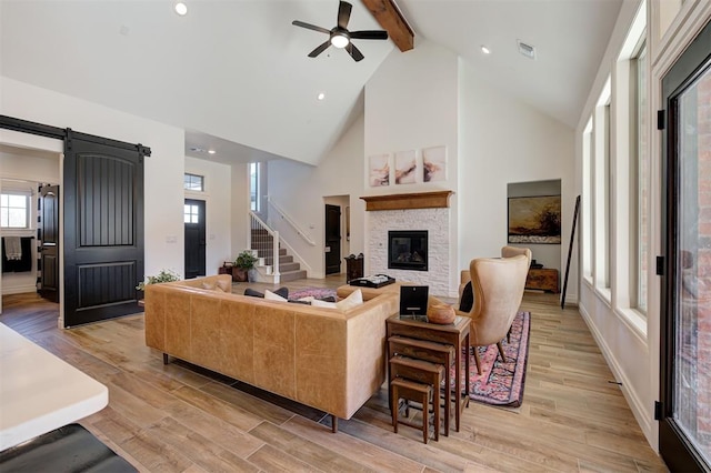 living room featuring a stone fireplace, high vaulted ceiling, beamed ceiling, a barn door, and light hardwood / wood-style floors