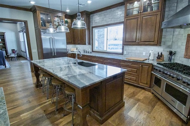 kitchen with wall chimney range hood, tasteful backsplash, an island with sink, sink, and double oven range