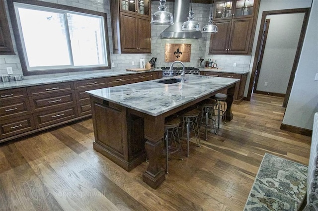 kitchen with light stone countertops, a kitchen island with sink, dark hardwood / wood-style floors, wall chimney range hood, and decorative backsplash