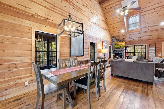 dining space featuring wood-type flooring, a fireplace, ceiling fan, wood walls, and high vaulted ceiling