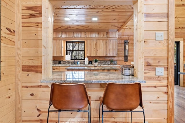 kitchen with wood walls, light brown cabinetry, wooden ceiling, and light stone counters