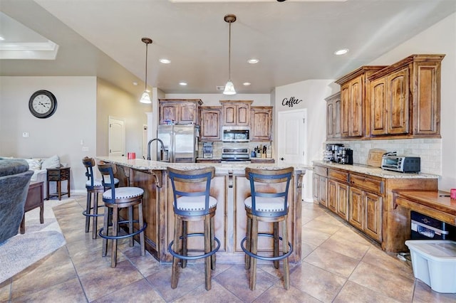 kitchen featuring a large island with sink, appliances with stainless steel finishes, decorative light fixtures, tasteful backsplash, and light stone counters