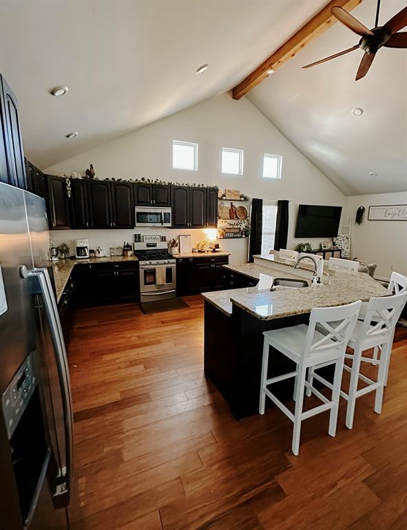 kitchen with light stone countertops, stainless steel appliances, an island with sink, beam ceiling, and a breakfast bar area