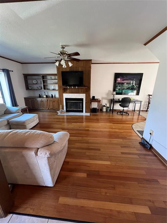 living area featuring a ceiling fan, wood finished floors, a textured ceiling, crown molding, and a fireplace