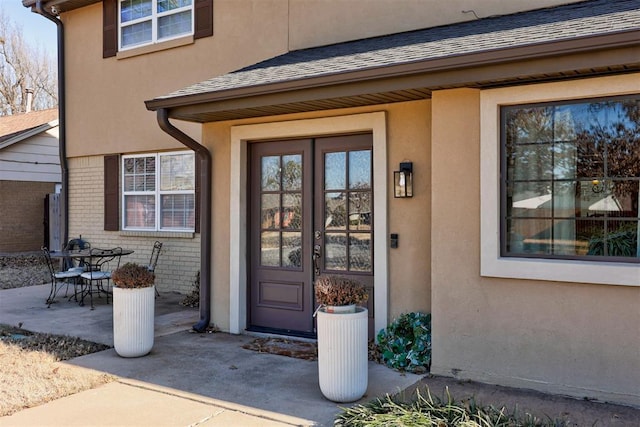 view of exterior entry featuring stucco siding, a shingled roof, outdoor dining area, brick siding, and a patio area