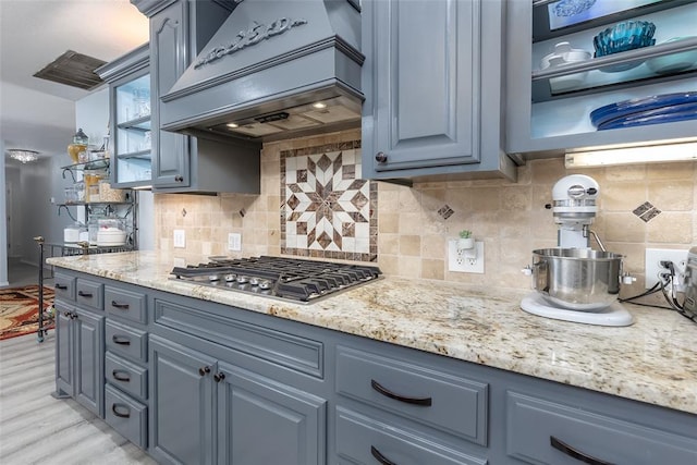 kitchen with stainless steel gas stovetop, light stone countertops, custom range hood, light wood-type flooring, and backsplash