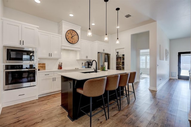 kitchen featuring pendant lighting, white cabinetry, stainless steel appliances, a center island with sink, and light wood-type flooring