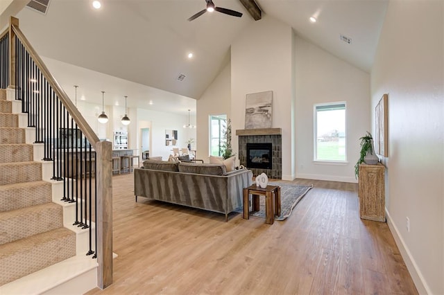 living room featuring ceiling fan, a tiled fireplace, light wood-type flooring, high vaulted ceiling, and beam ceiling