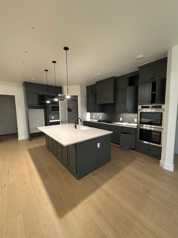 kitchen with sink, an island with sink, decorative backsplash, decorative light fixtures, and light wood-type flooring