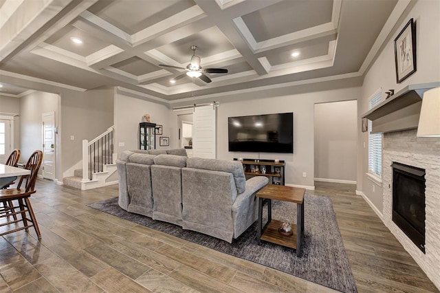 living room featuring ceiling fan, coffered ceiling, ornamental molding, a stone fireplace, and a barn door