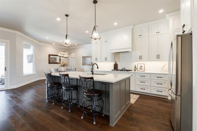 kitchen with custom range hood, stainless steel refrigerator, hanging light fixtures, white cabinets, and an island with sink