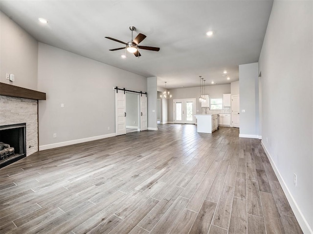 unfurnished living room with a barn door, light wood-type flooring, a stone fireplace, and ceiling fan with notable chandelier