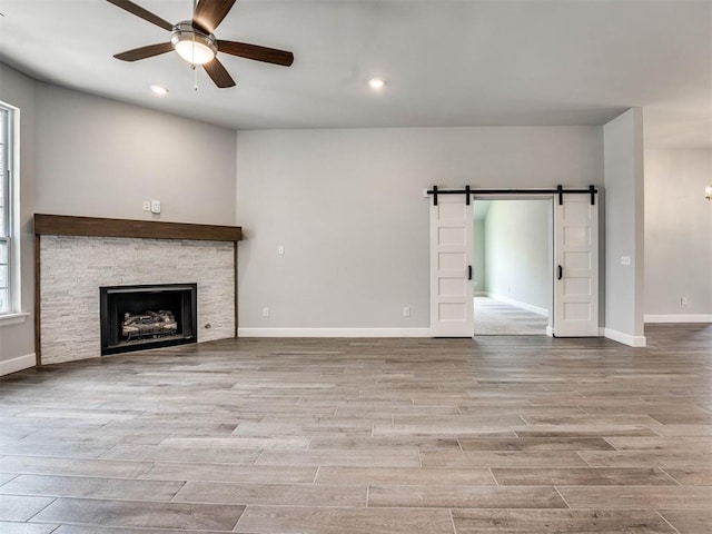 unfurnished living room with ceiling fan, a barn door, a stone fireplace, and a healthy amount of sunlight