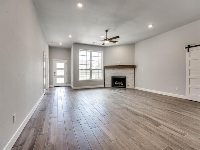 unfurnished living room with ceiling fan, light hardwood / wood-style floors, a stone fireplace, and a barn door