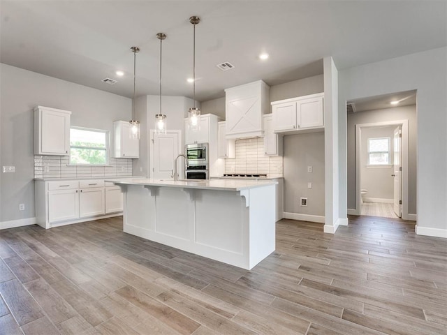 kitchen with an island with sink, stainless steel microwave, tasteful backsplash, pendant lighting, and white cabinets