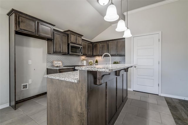 kitchen featuring decorative light fixtures, decorative backsplash, a kitchen island with sink, dark brown cabinetry, and light stone countertops