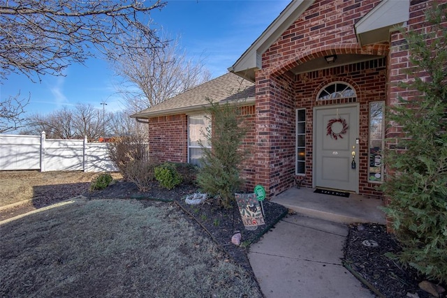 view of exterior entry with a shingled roof, fence, and brick siding