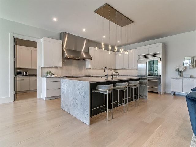 kitchen with pendant lighting, wall chimney exhaust hood, light hardwood / wood-style flooring, white cabinetry, and a center island with sink