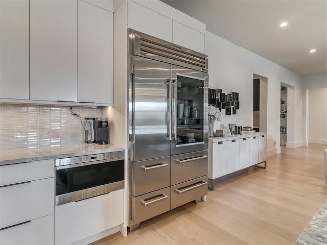 kitchen with white cabinets, stainless steel built in fridge, tasteful backsplash, light stone counters, and light hardwood / wood-style flooring