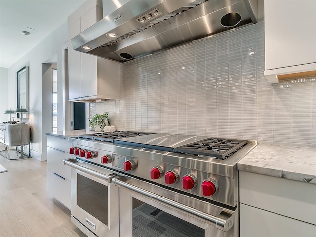 kitchen featuring tasteful backsplash, wall chimney range hood, white cabinetry, light wood-type flooring, and range with two ovens