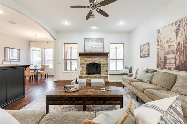 living room featuring crown molding, dark hardwood / wood-style floors, ceiling fan with notable chandelier, and a stone fireplace