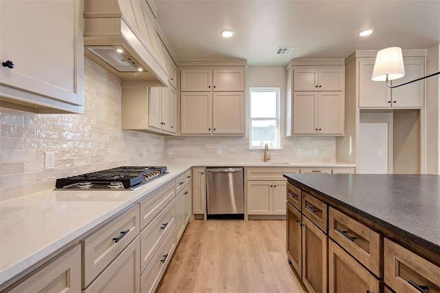 kitchen with appliances with stainless steel finishes, decorative light fixtures, sink, light wood-type flooring, and custom range hood