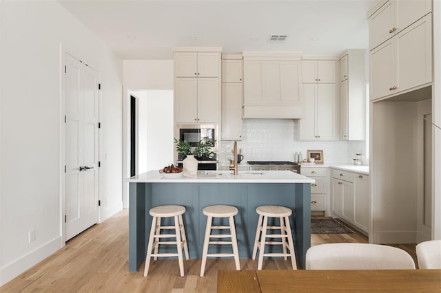 kitchen with wall chimney exhaust hood, sink, backsplash, a kitchen island with sink, and light wood-type flooring