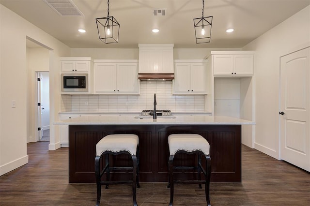 kitchen featuring hanging light fixtures, white cabinets, a kitchen island with sink, and black microwave